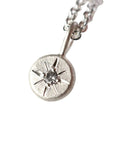 A sterling silver pendant necklace featuring a textured circular disc with a starburst design and a central clear gemstone, resting on a white textured stone surface.