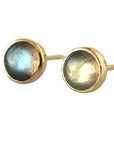 A pair of gold stud earrings with round iridescent labradorite stones, resting on a reflective light gray surface.