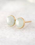 A pair of gold stud earrings with round, pale greenish-white cabochon gemstones, resting on a soft, light-colored textured surface.