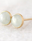 A pair of gold stud earrings with round, pale greenish-white cabochon gemstones, resting on a soft, light-colored textured surface.