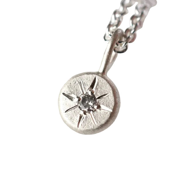 A sterling silver pendant necklace featuring a textured circular disc with a starburst design and a central clear gemstone, resting on a white textured stone surface.