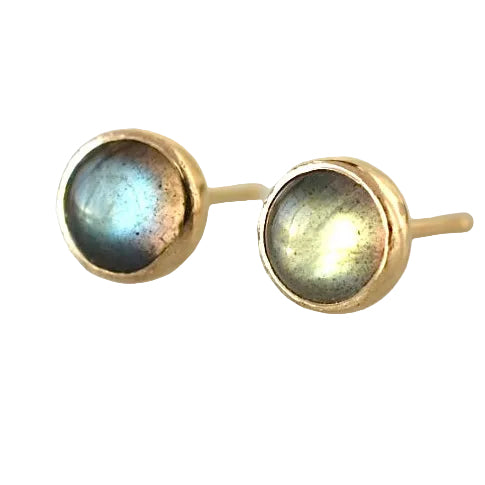 A pair of gold stud earrings with round iridescent labradorite stones, resting on a reflective light gray surface.