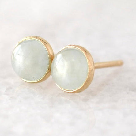 A pair of gold stud earrings with round, pale greenish-white cabochon gemstones, resting on a soft, light-colored textured surface.