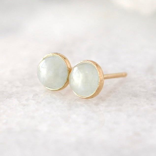 A pair of gold stud earrings with round, pale greenish-white cabochon gemstones, resting on a soft, light-colored textured surface.